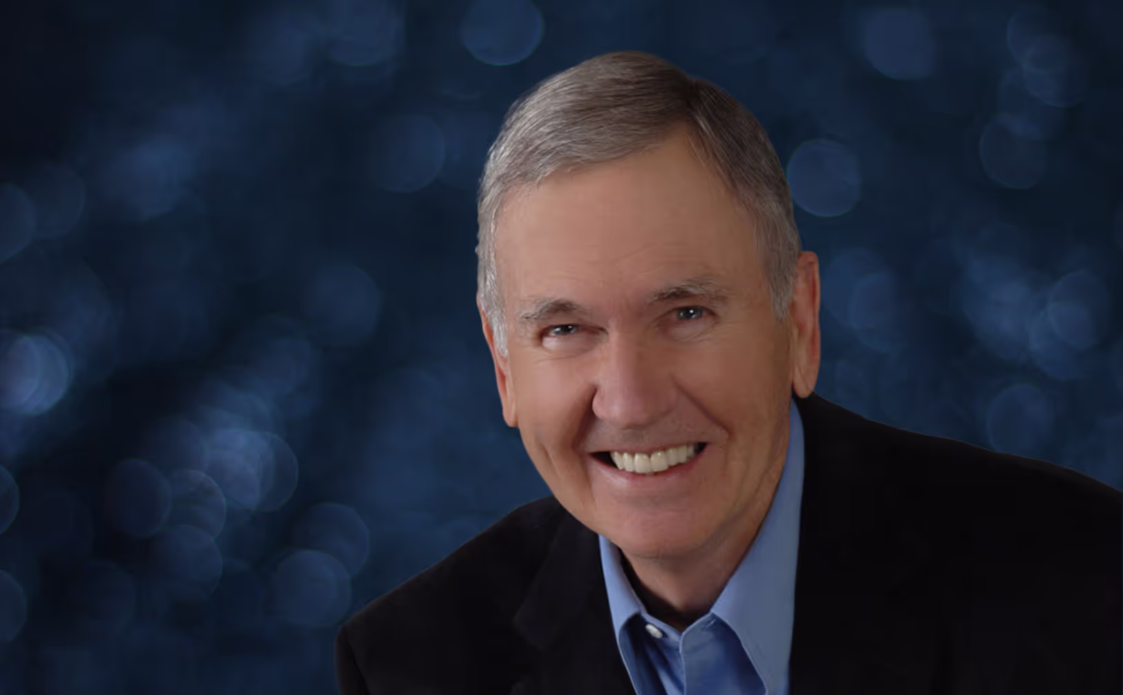 A man wearing a blue shirt and a black jacket smiling.